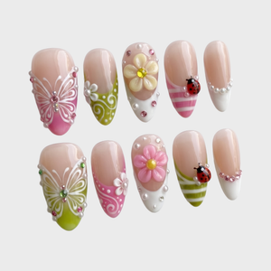 A set of glossy press-on nails decorated with 3D pink and yellow flowers, red ladybugs, white butterflies, and pearls on pink and green backgrounds.