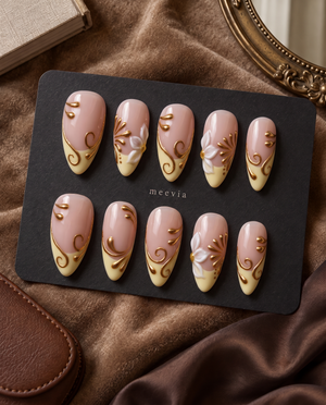 Glossy nude and cream press-on nails with white floral accents and raised gold swirls