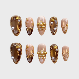 A set of press-on nails featuring brown and nude bases decorated with gold filigree, pearl accents, and metallic beads.