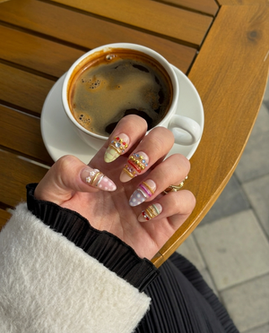 A set of ten nude press-on nails decorated with colorful rhinestones, gold chains, silver bands, white flower charms, and pastel patterns.