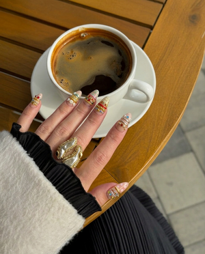 A set of ten nude press-on nails decorated with colorful rhinestones, gold chains, silver bands, white flower charms, and pastel patterns.