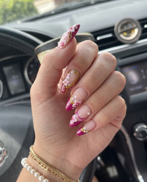 A set of magenta glitter French tip press-on nails decorated with 3D floral designs, gold filigree, and small white pearls.