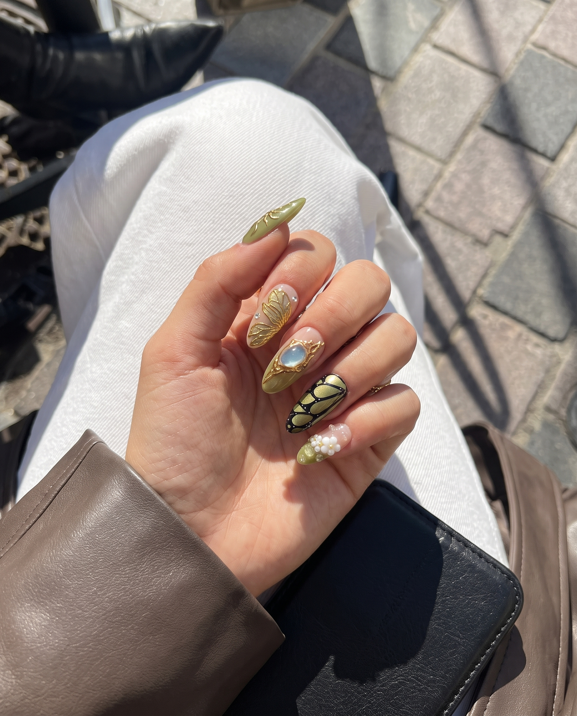 A set of press-on nails featuring olive green and pink bases decorated with gold butterfly wings, 3D flowers, and a large gemstone.