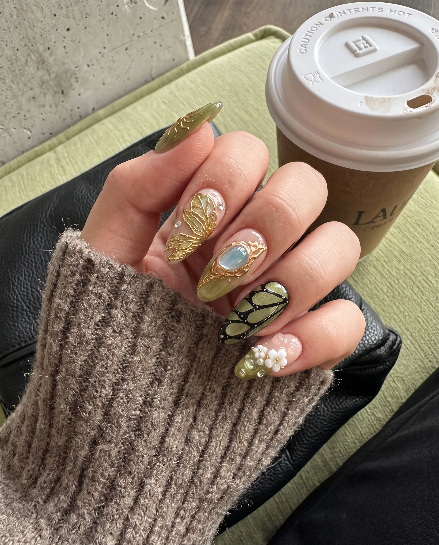 A set of press-on nails featuring olive green and pink bases decorated with gold butterfly wings, 3D flowers, and a large gemstone.