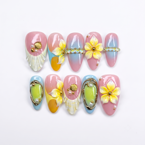 3D pastel press-on nails featuring yellow flowers, white seashells, gold accents, and a pink and blue gradient background.