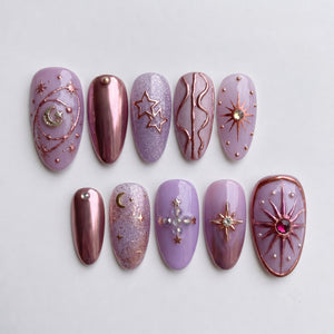 Lavender press-on nails decorated with metallic rose gold celestial designs, stars, moons, and rhinestones.