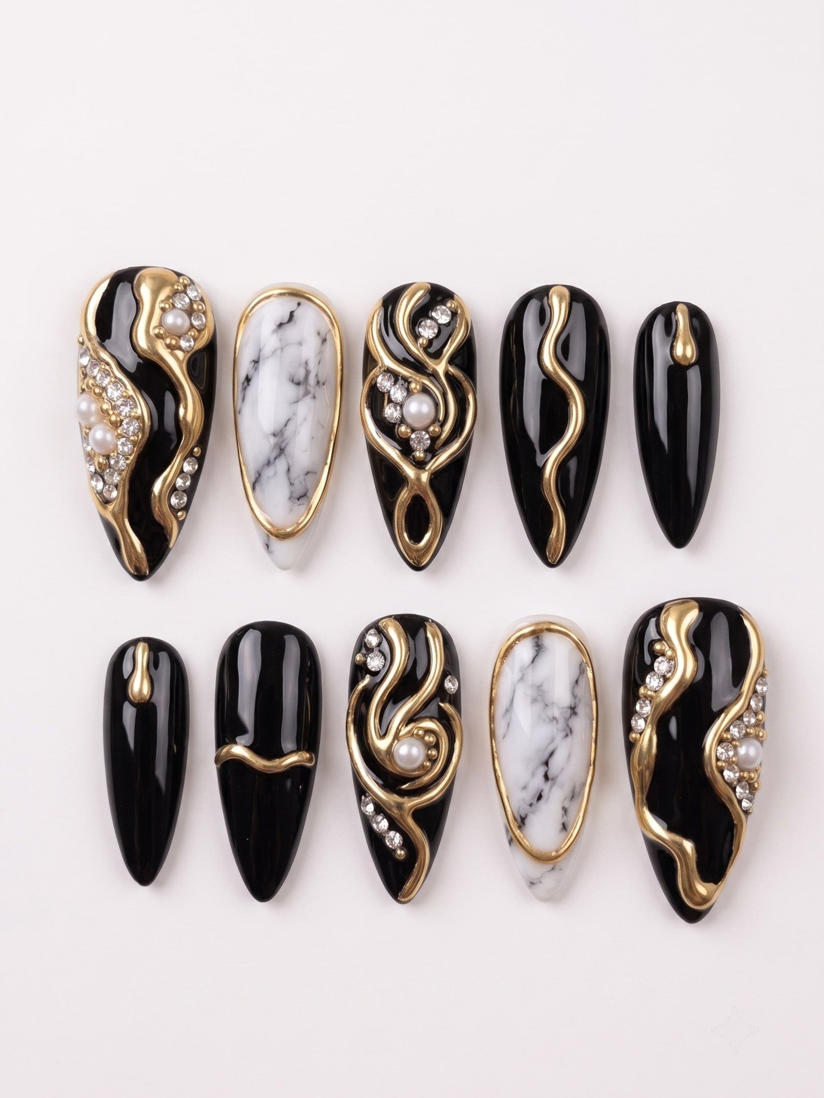 Black press-on nails with white marble patterns, gold swirls, pearls, and rhinestones.