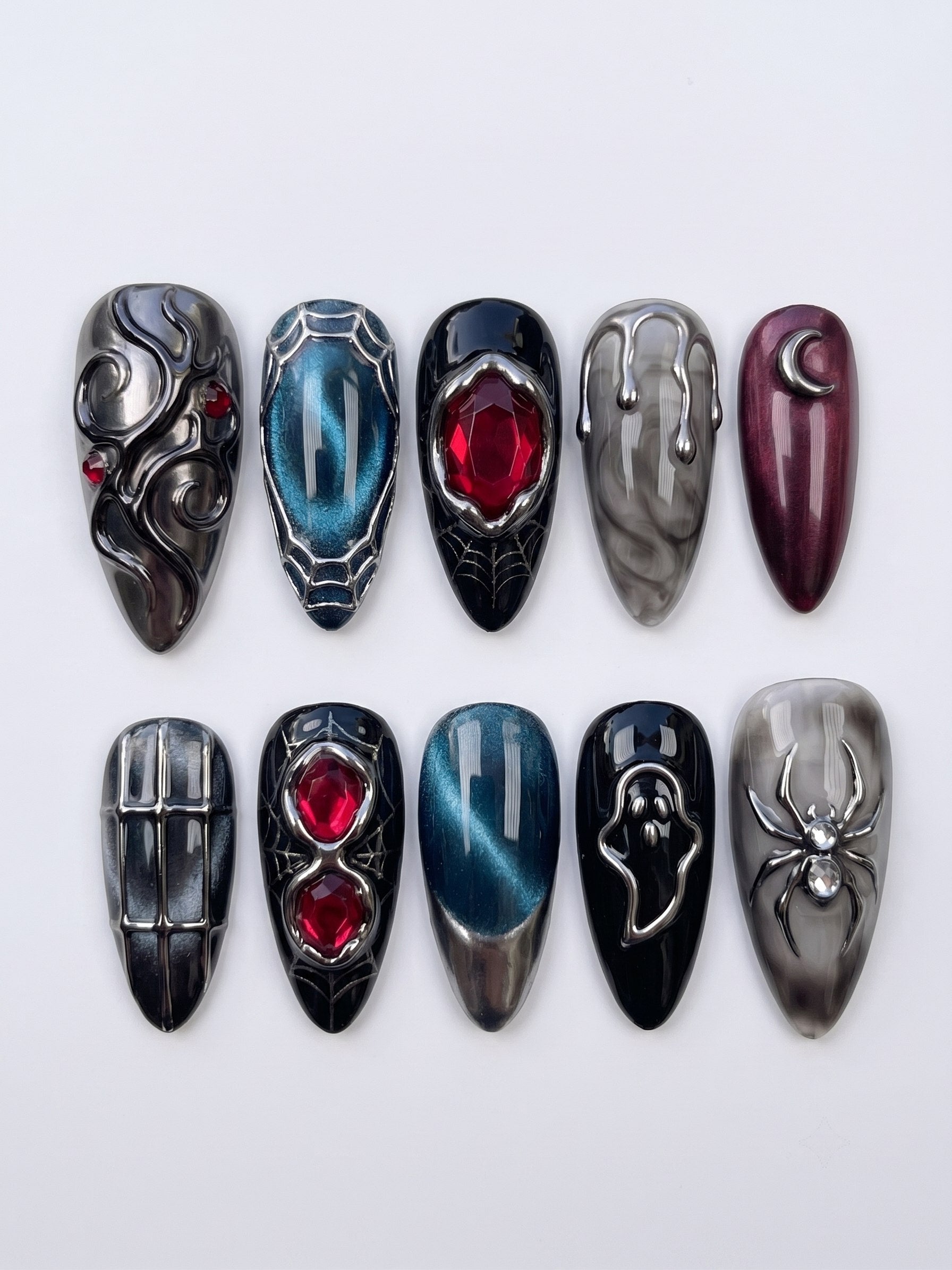 A set of gothic press-on nails featuring black, grey marble, and teal cat-eye bases with silver metallic spider, ghost, crescent moon, and red gemstone details.