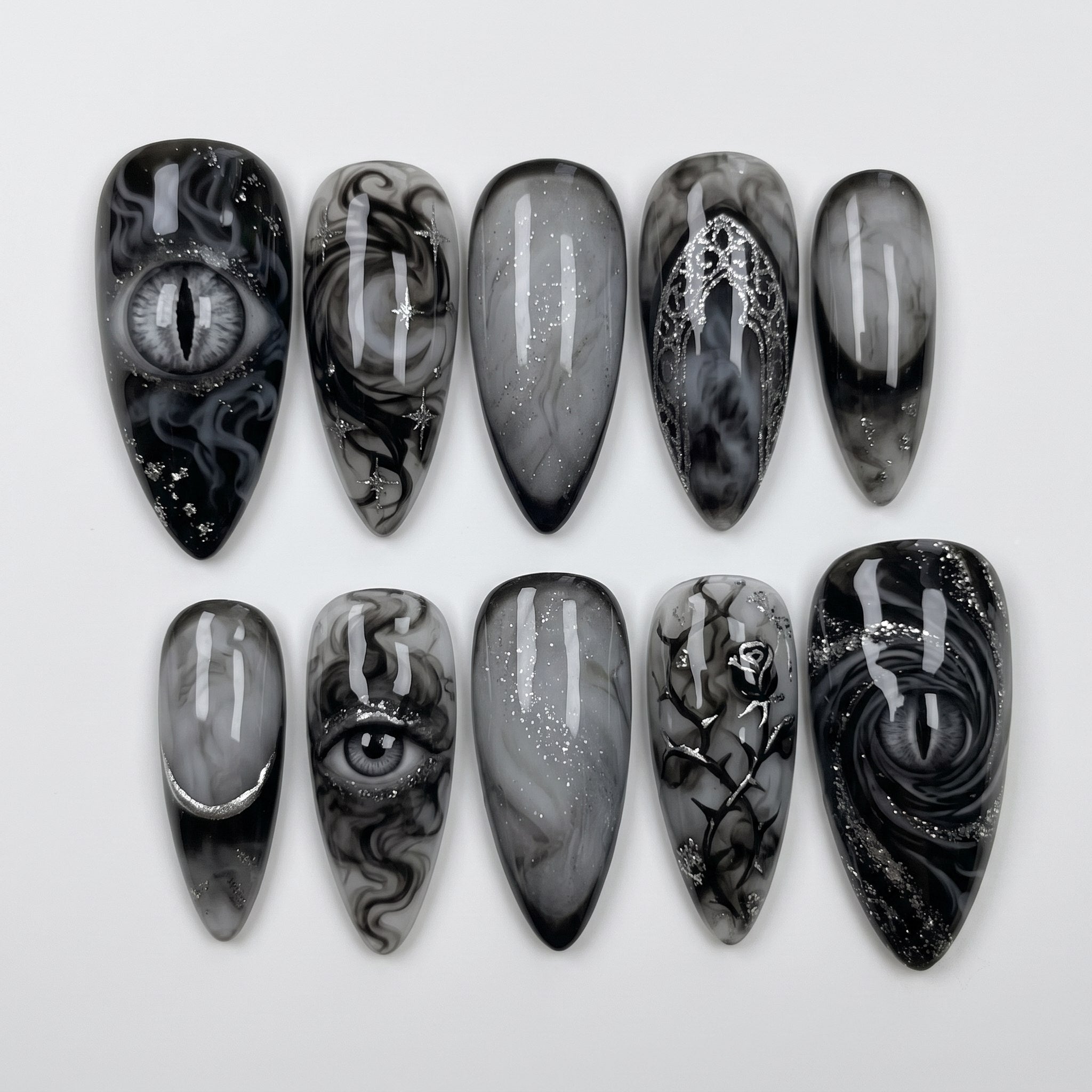 A set of gothic press-on nails featuring black and grey smoky marble designs, detailed eye motifs, and silver metallic rose, moon, and arch details.