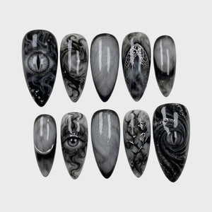 A set of gothic press-on nails featuring black and grey smoky marble designs, detailed eye motifs, and silver metallic rose, moon, and arch details.