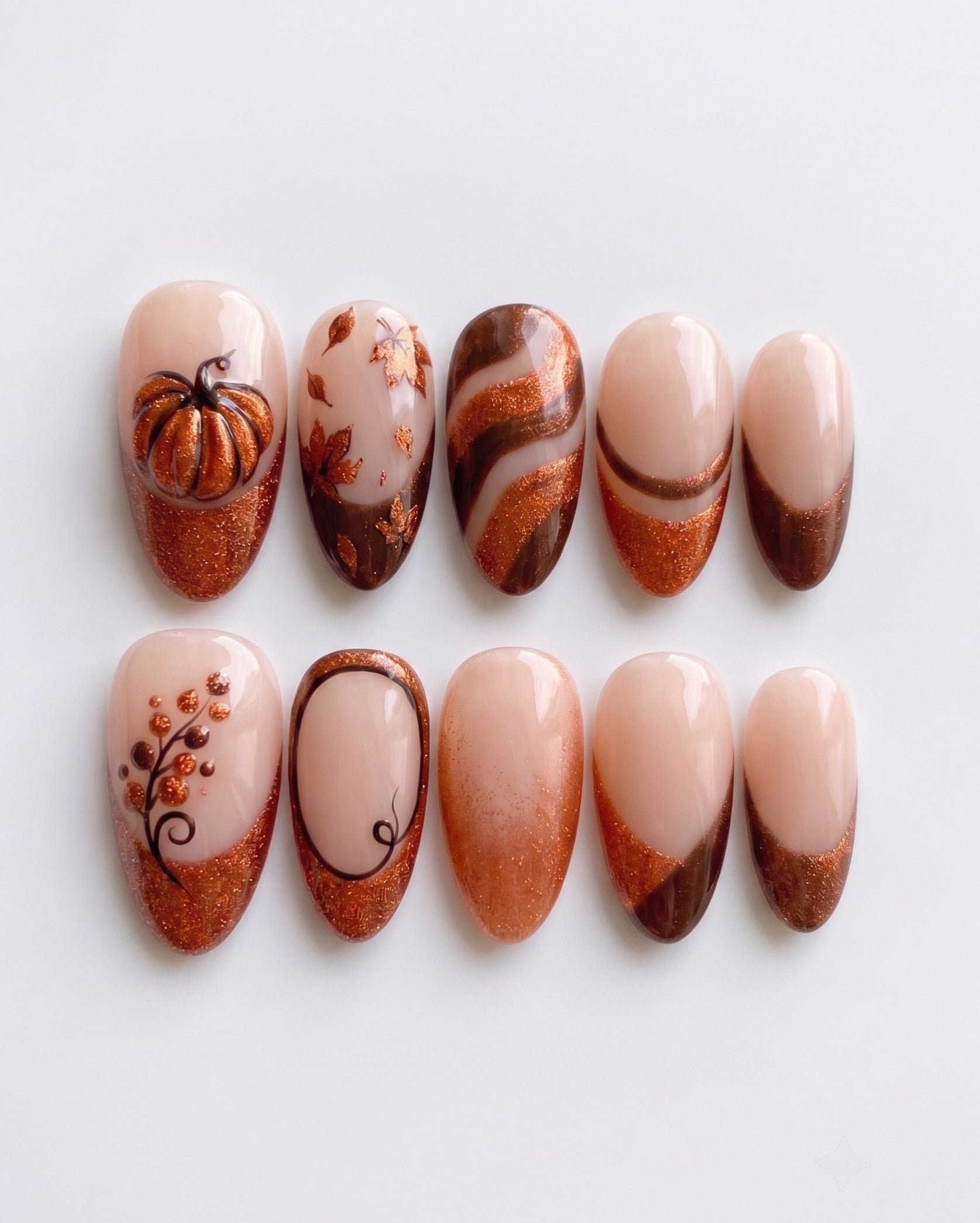 A set of autumn-themed press-on nails featuring copper glitter French tips, a 3D metallic pumpkin, fall leaves, and brown swirl designs on a nude base.