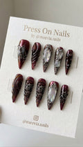 Stiletto-shaped burgundy press-on nails with 3D silver accents and iridescent pearl details.