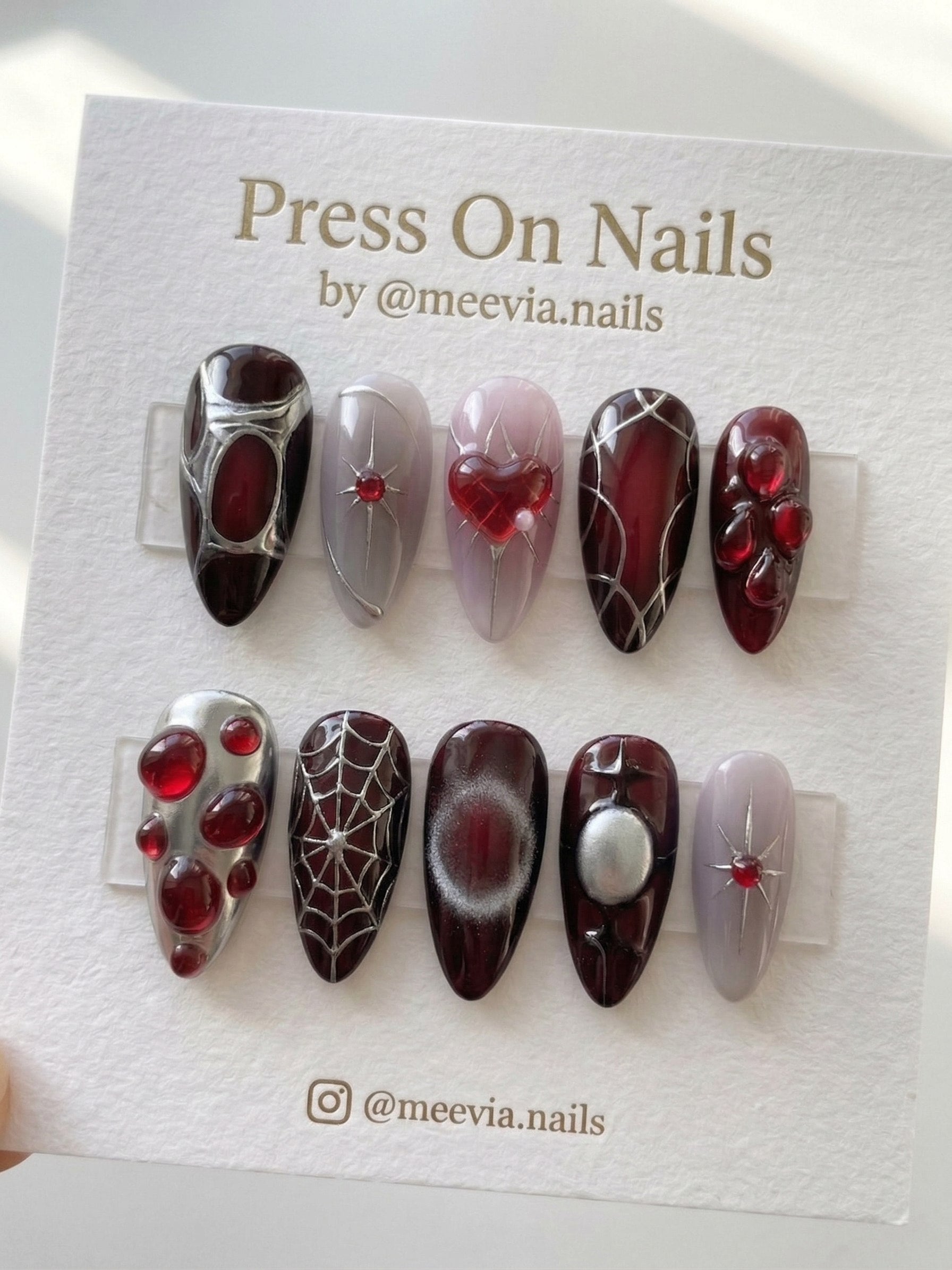 Almond-shaped gothic press-on nail set with deep red base, silver spiderwebs, 3D red heart, and metallic star motifs.