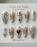 Almond-shaped white and nude press-on nails with silver 3D swirls, a red heart, and a gothic cross.