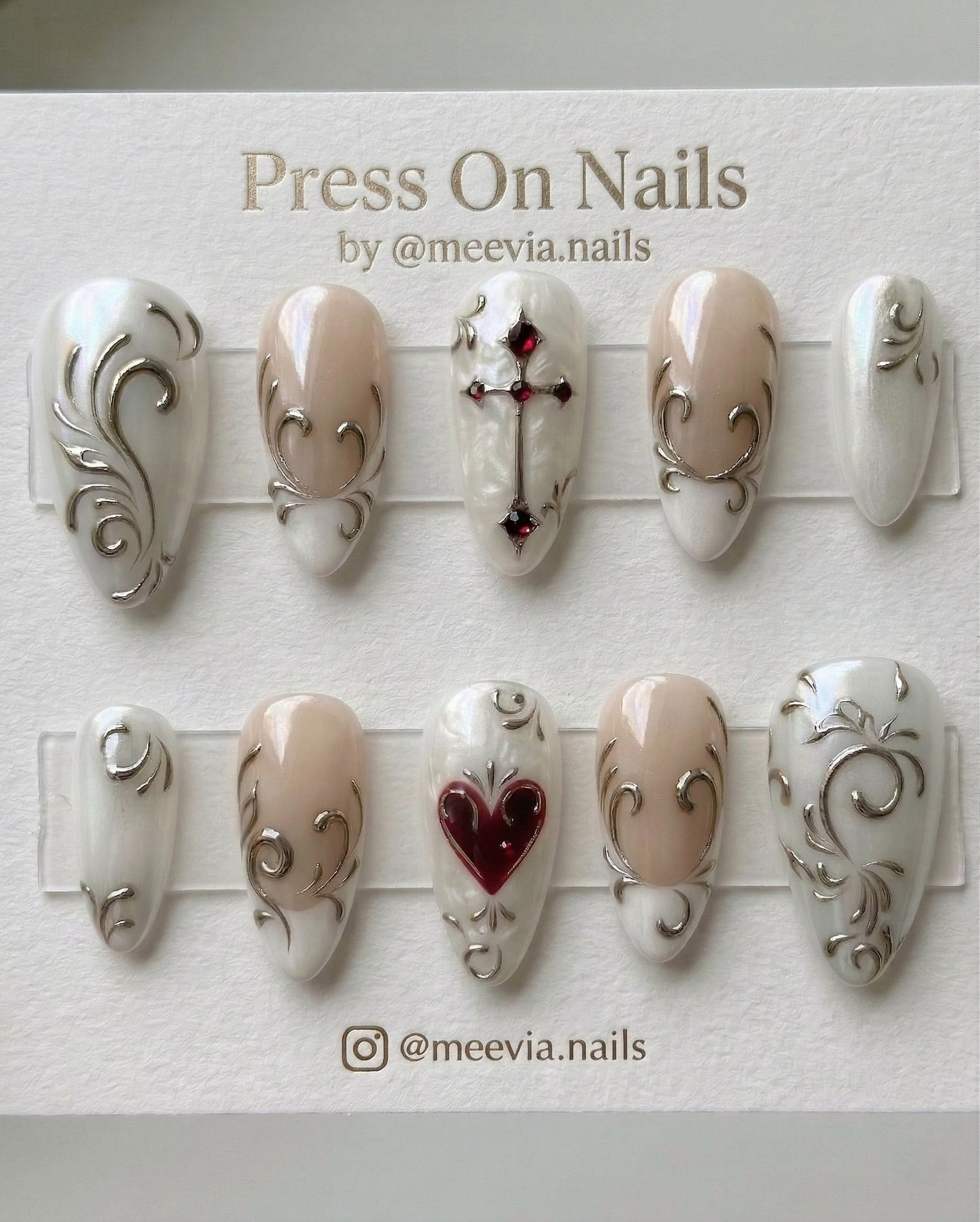 Almond-shaped white and nude press-on nails with silver 3D swirls, a red heart, and a gothic cross.