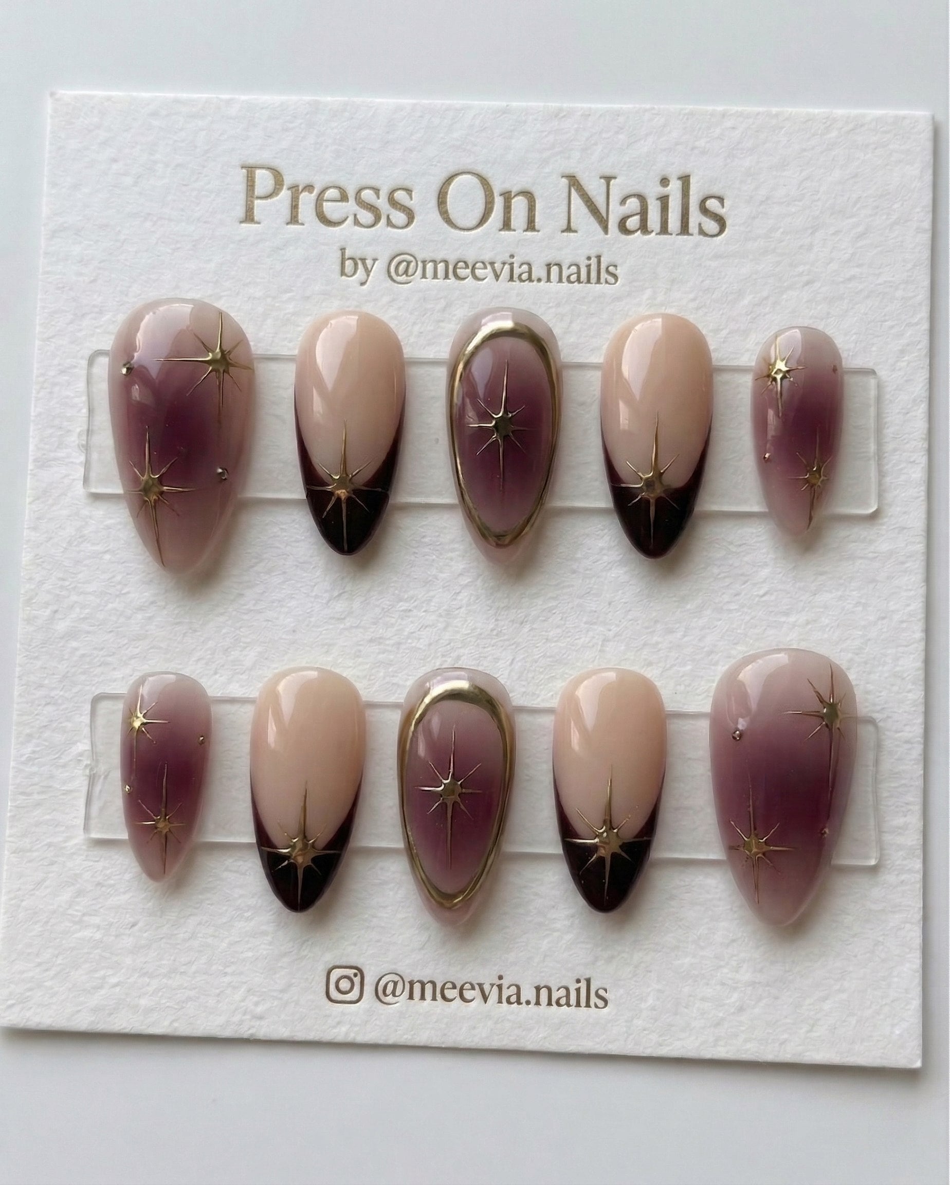 Almond-shaped plum and nude press-on nail set with gold celestial star motifs and metallic gold frames.