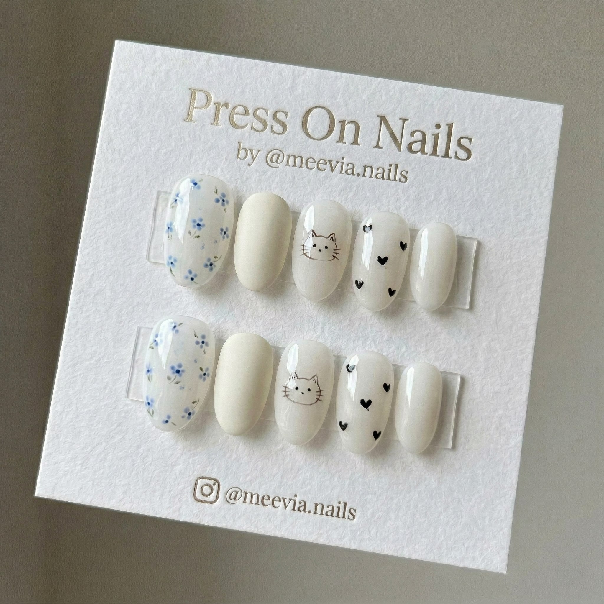 Oval white press-on nail set with blue floral patterns, a cute cat face, and black hearts