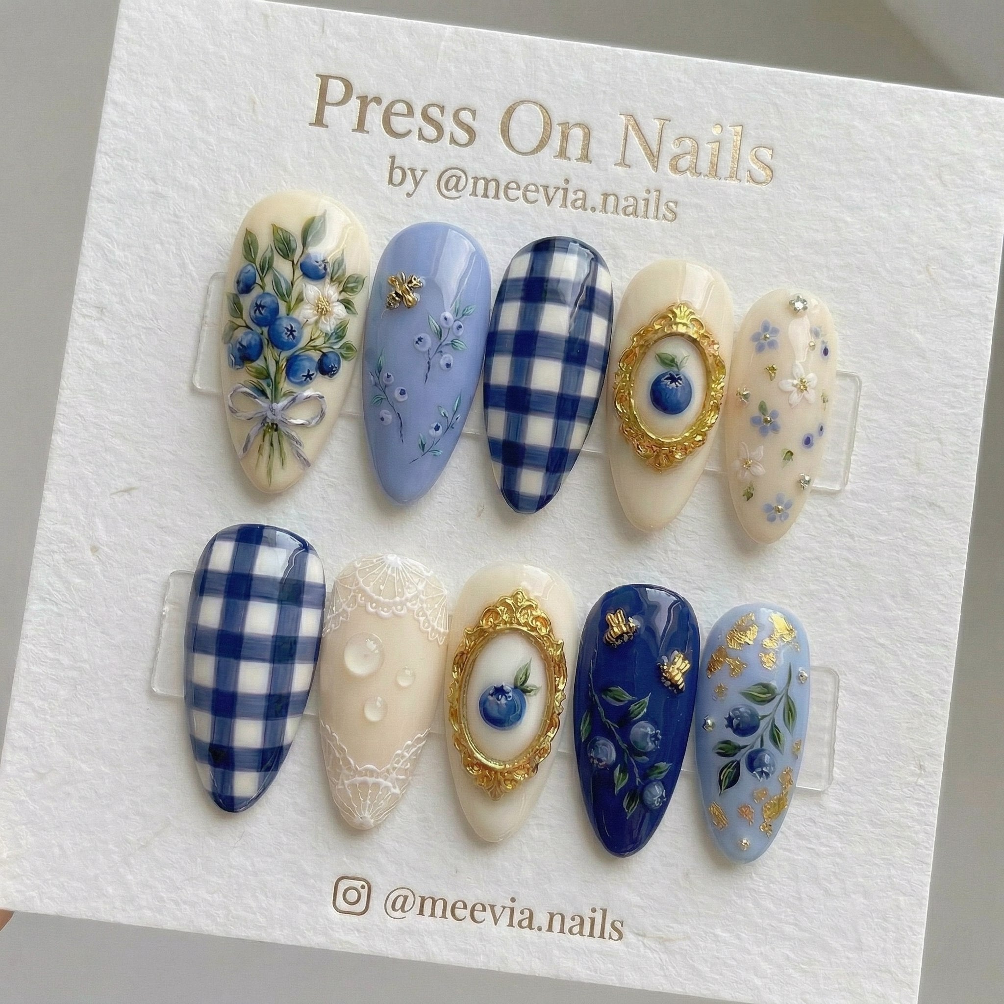 Almond-shaped blue and cream press-on nail set featuring blueberries, gingham patterns, and 3D gold bee accents.