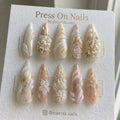 Stiletto-shaped press-on nail set with 3D white flowers, lace patterns, gold foil, and pearls on a soft pink base.
