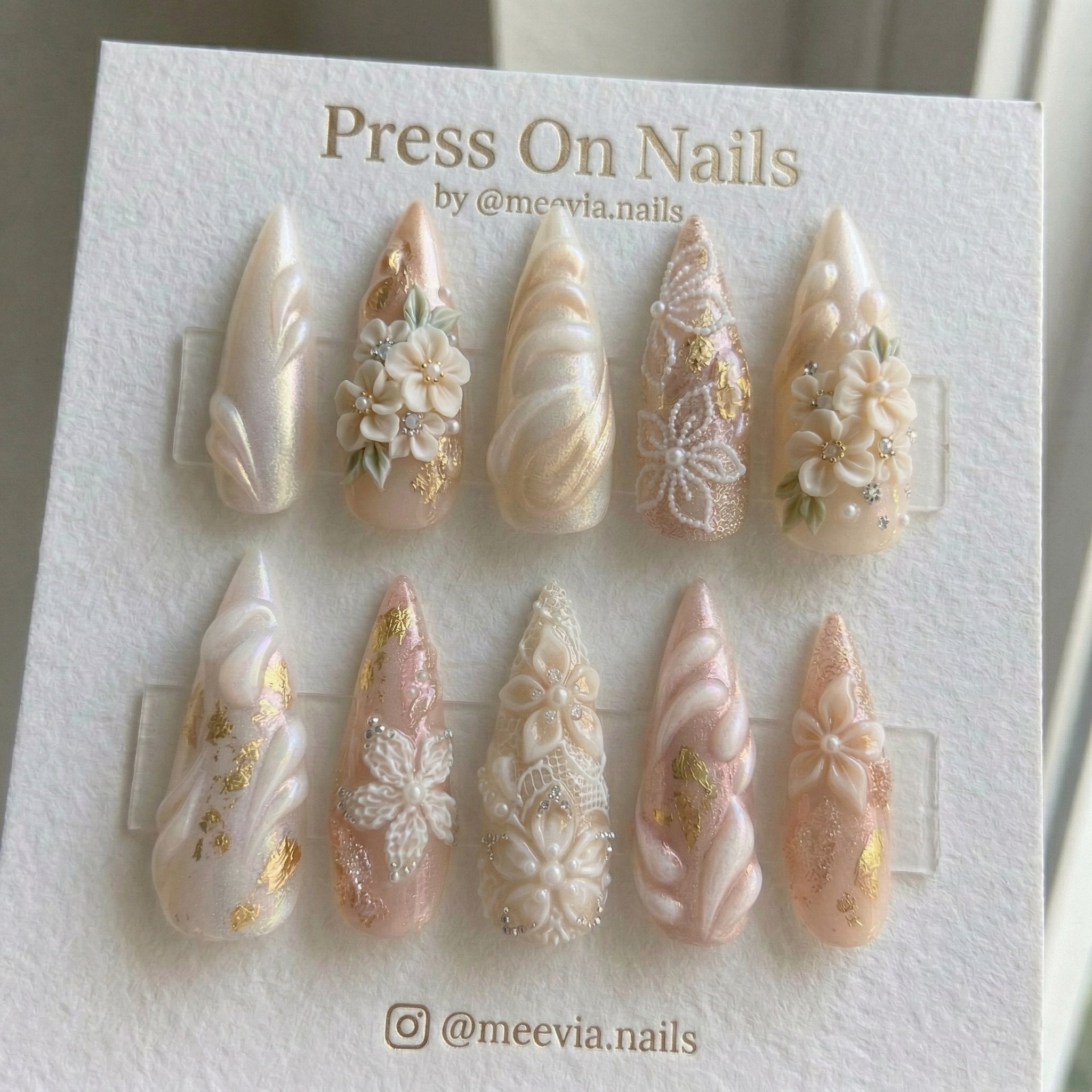 Stiletto-shaped press-on nail set with 3D white flowers, lace patterns, gold foil, and pearls on a soft pink base.