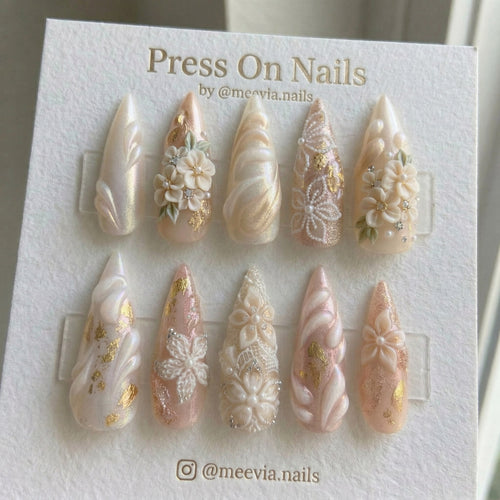 Stiletto-shaped press-on nail set with 3D white flowers, lace patterns, gold foil, and pearls on a soft pink base.