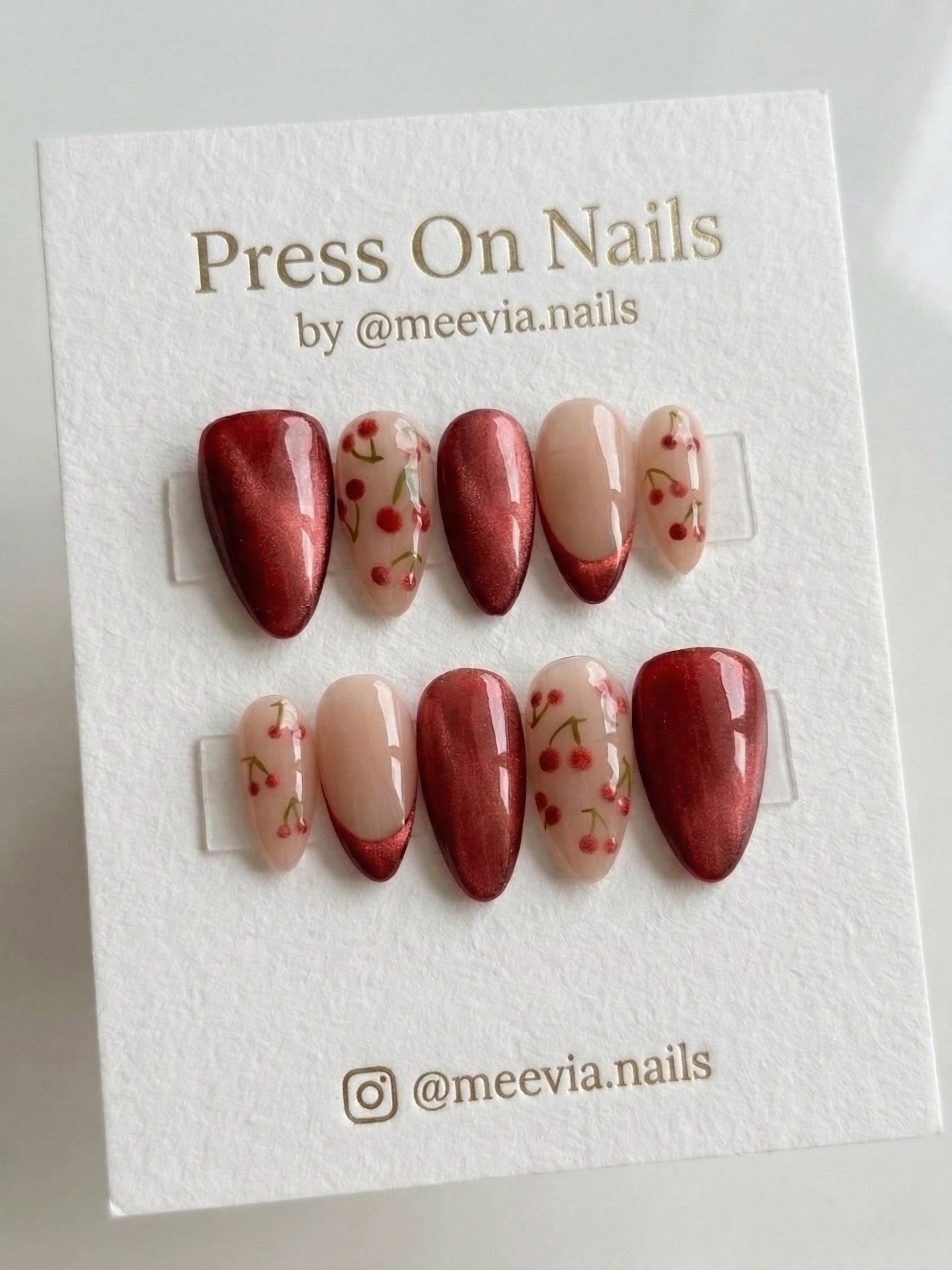 Almond-shaped press-on nail set featuring red shimmer finish and hand-painted cherry motifs on a nude base.