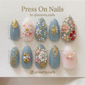 Almond-shaped dusty blue press-on nails with 3D white flowers, gold vines, and pearl embellishments.