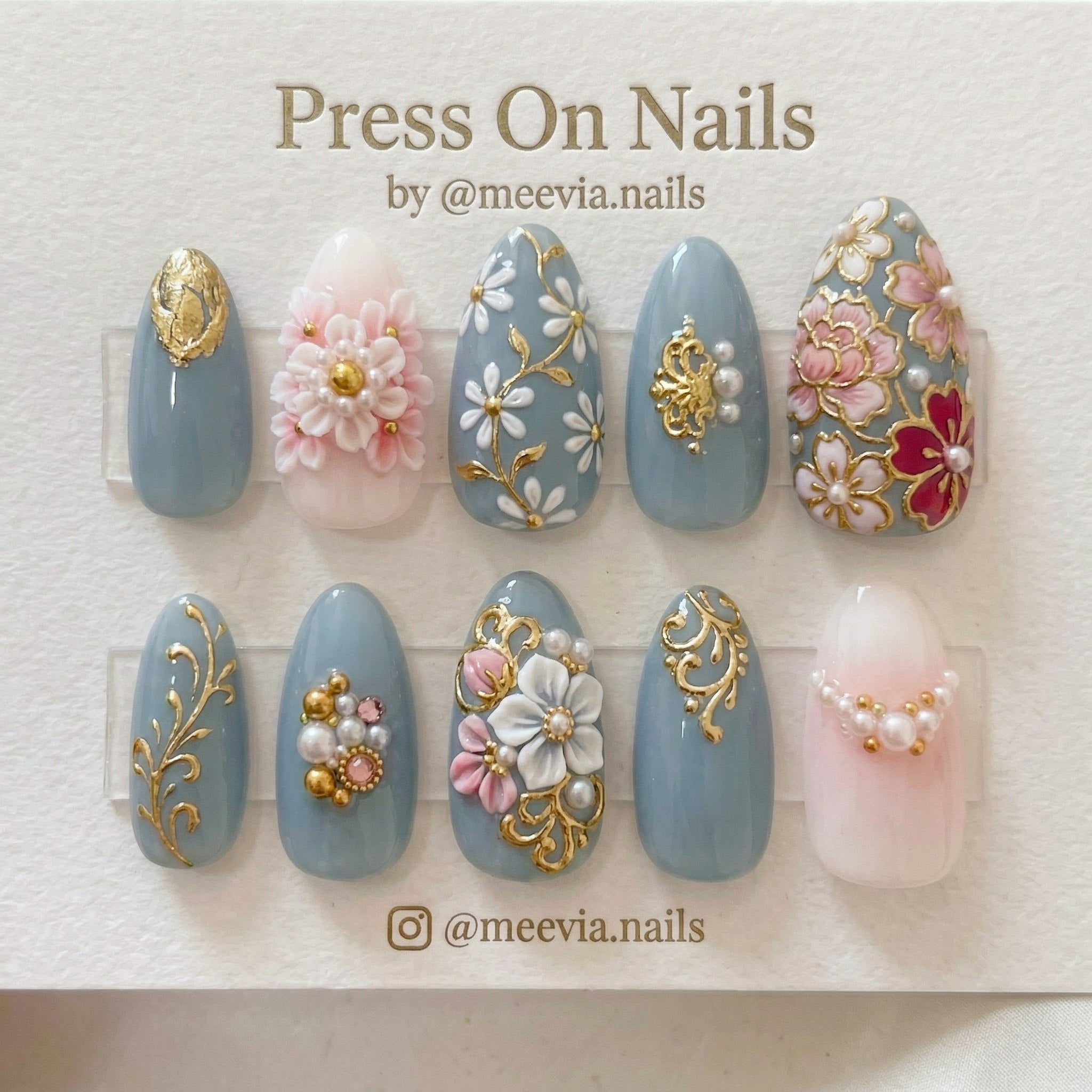 Almond-shaped dusty blue press-on nails with 3D white flowers, gold vines, and pearl embellishments.