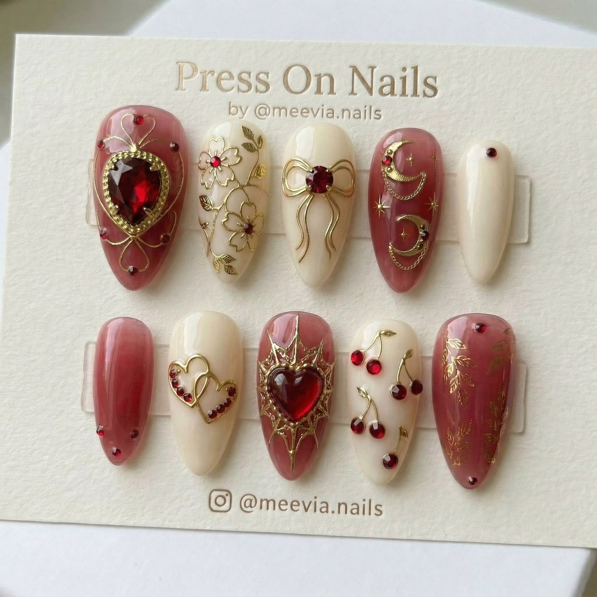 Almond-shaped press on nails with red heart gems, gold celestial moons, and cherry motifs on ivory and jelly red bases.