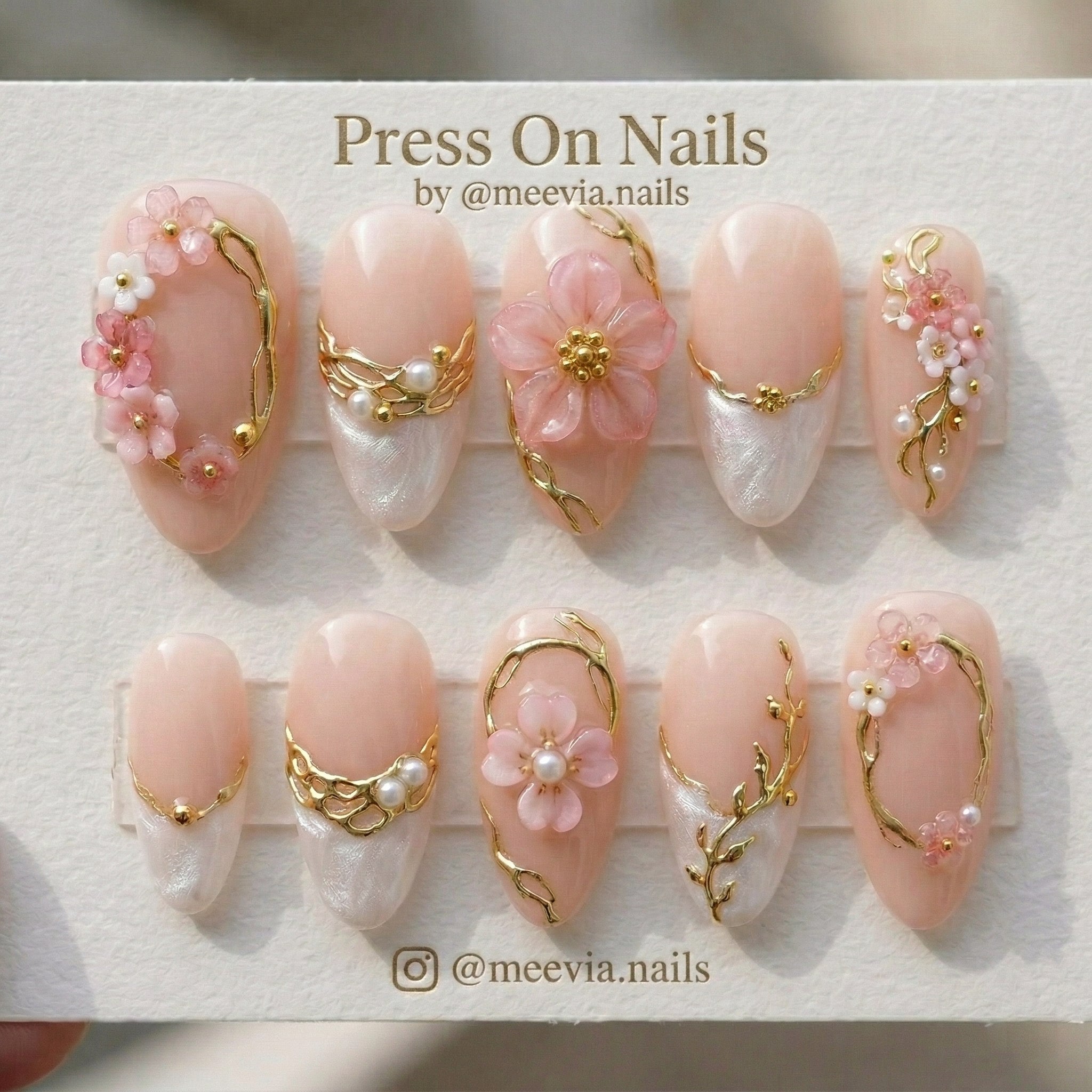 Almond-shaped blush press-on nails with 3D pink flowers, gold vines, and shimmering white tips.