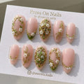 Almond-shaped pink press-on nails with 3D gold frames, white flowers, and green leaves