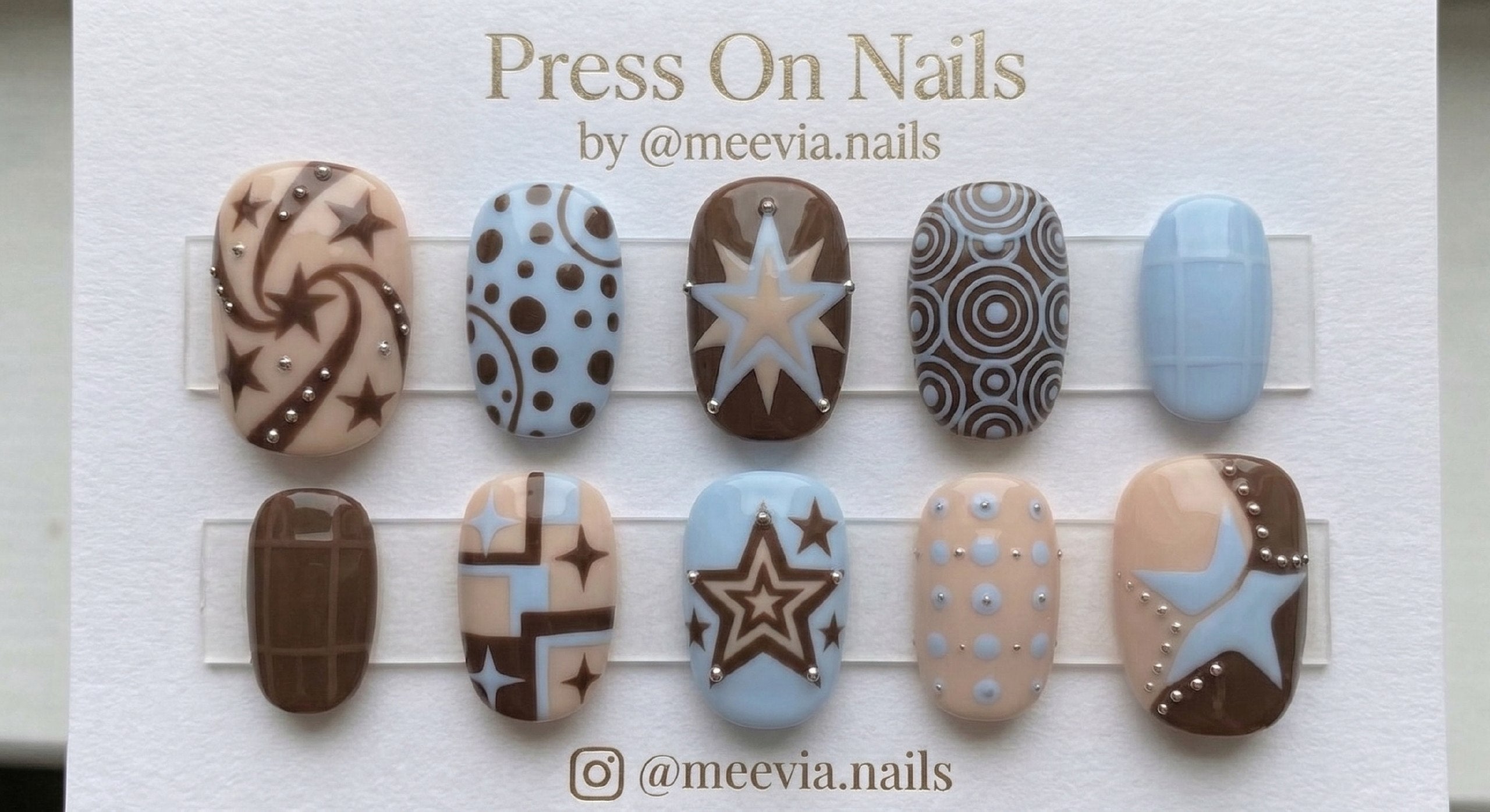Short rounded press-on nail set featuring stars, circles, and grid patterns in brown, tan, and light blue with silver stud accents.