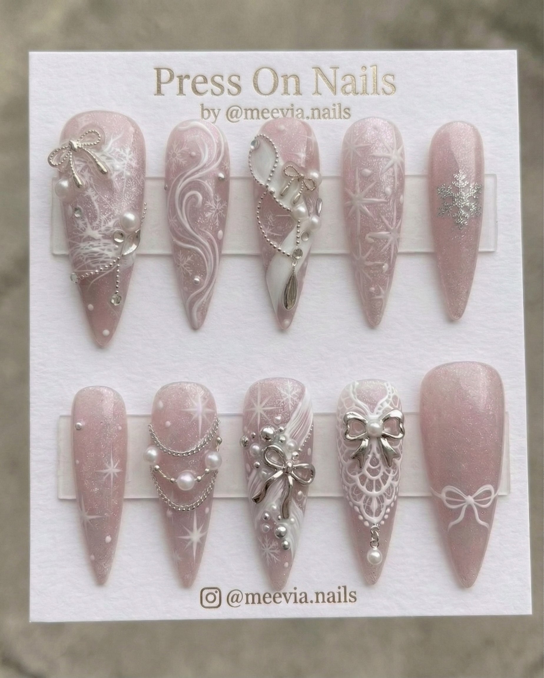 Long stiletto press-on nails with pink shimmer, 3D silver bows, and white snowflake nail art.