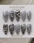 Long stiletto press-on nail set with grey cat-eye shimmer, 3D silver chrome stars, and metallic heart motifs.