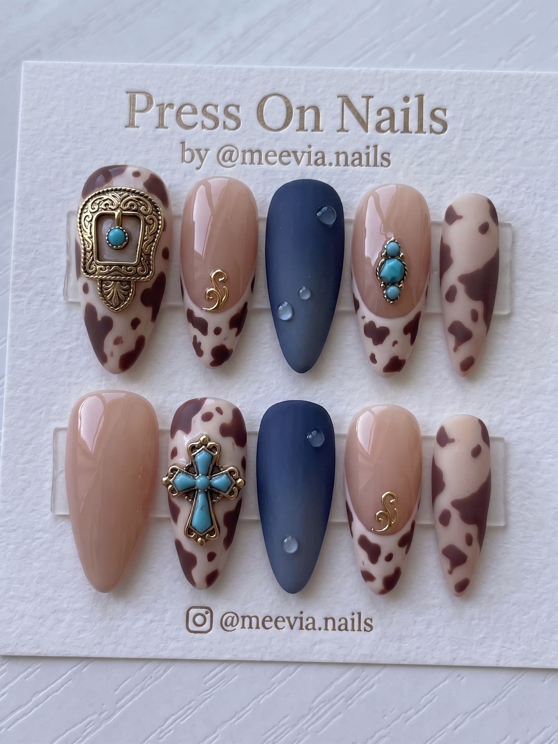 Almond-shaped western press-on nails featuring brown cow print, turquoise charms, and matte navy blue with 3D droplets.