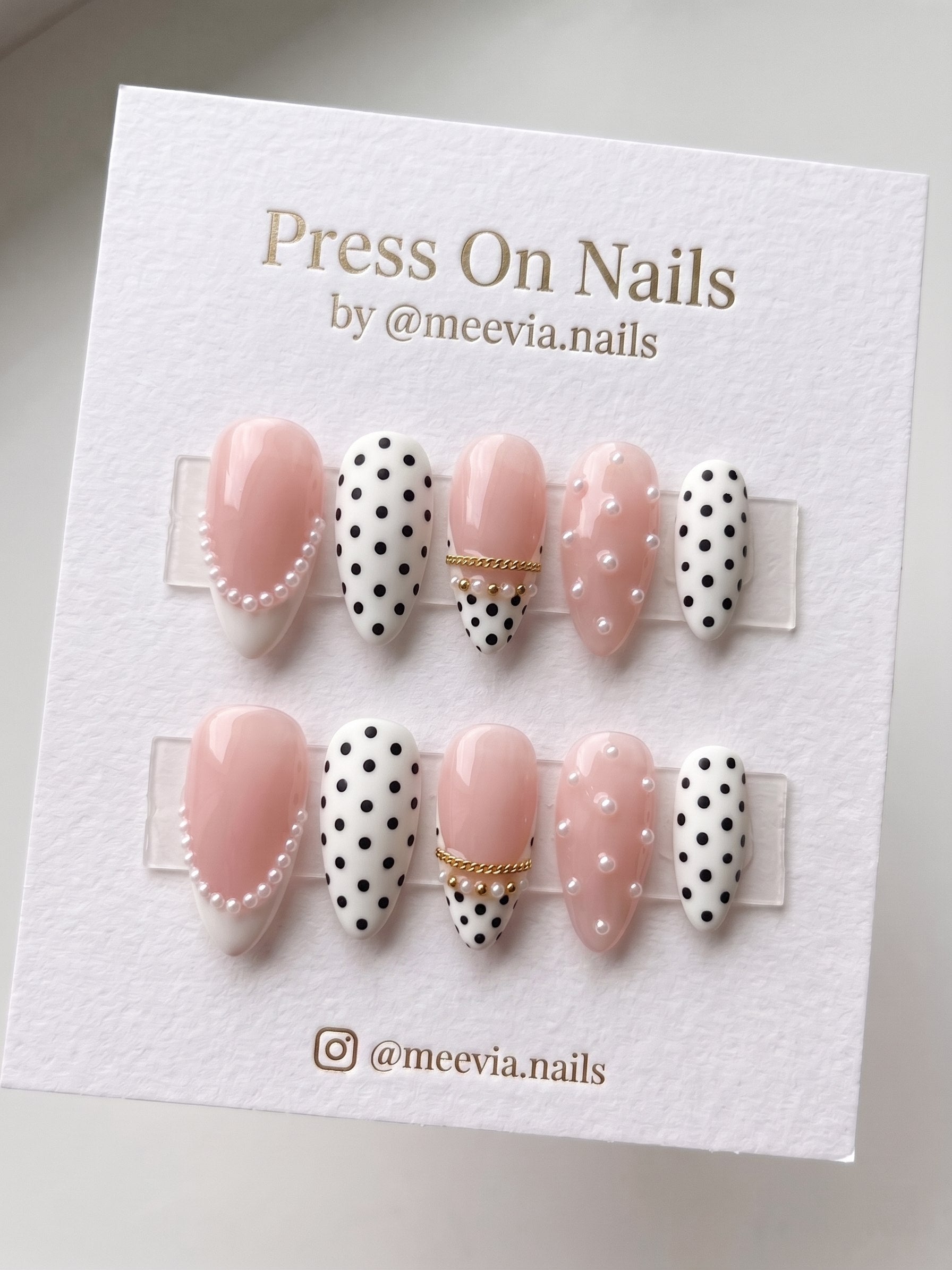 Almond-shaped pink press on nails with polka dots, pearls, and gold chain accents.