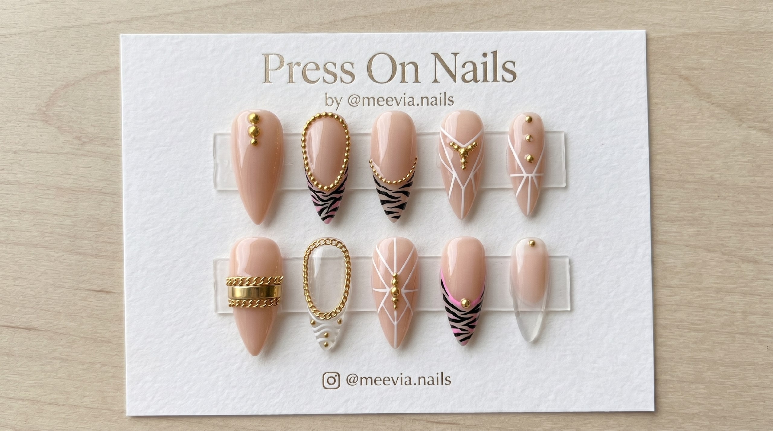 Stiletto-shaped nude press-on nails featuring zebra print, gold chains, and 3D stud nail art details.