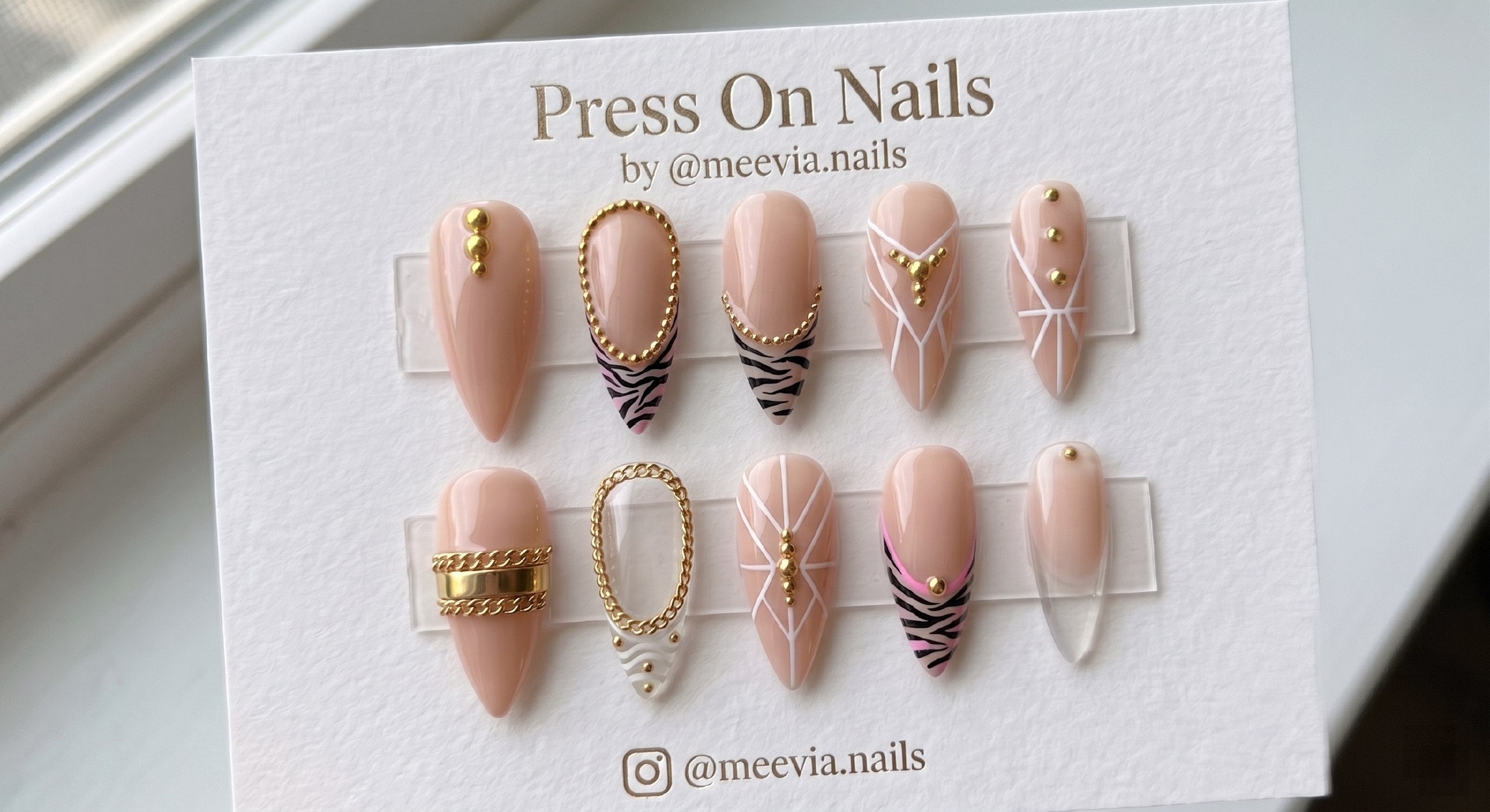 Stiletto-shaped nude press-on nails with black zebra print, gold chain embellishments, and white geometric lines.