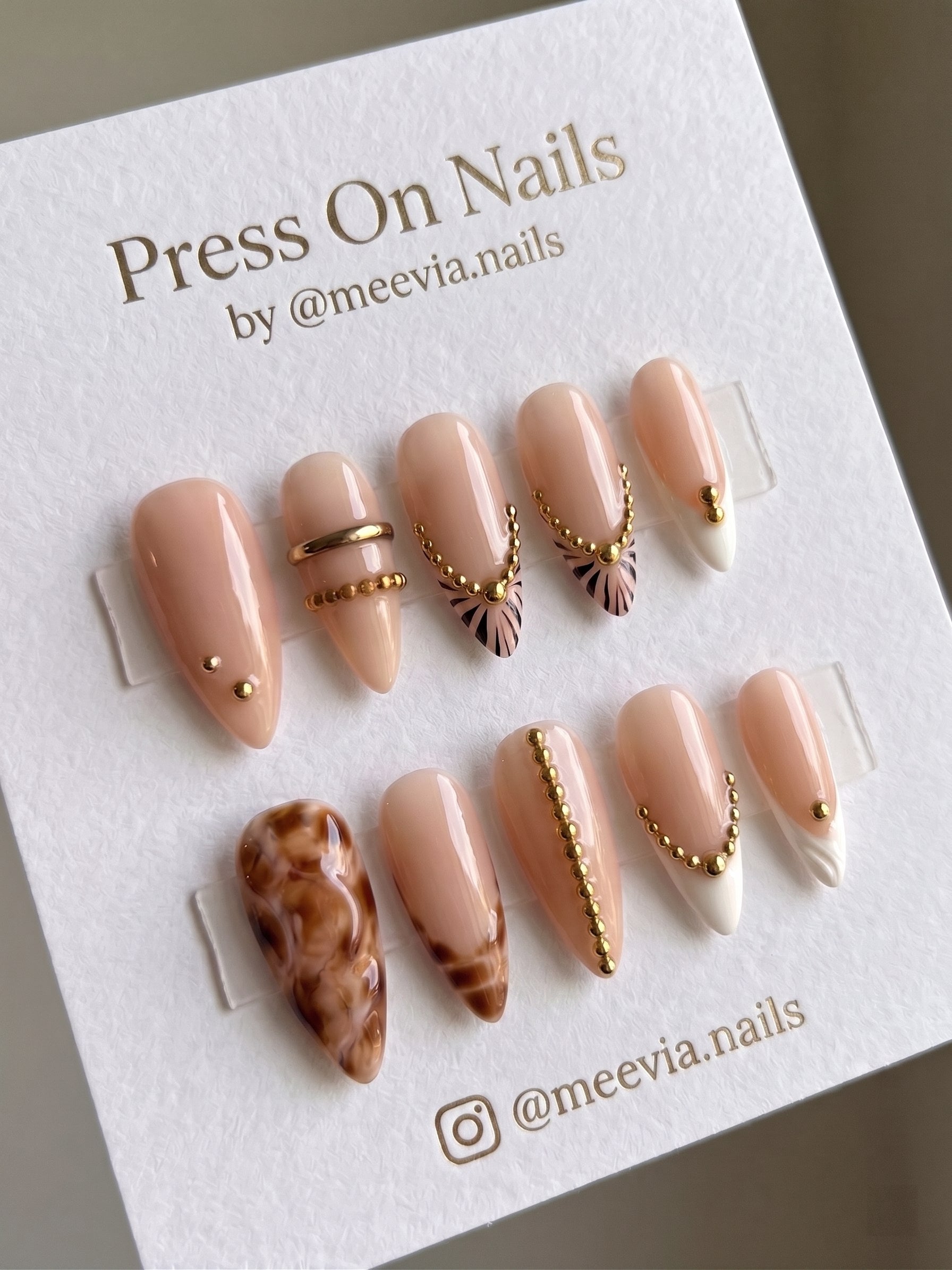 Almond-shaped nude press-on nails with gold bead chain charms and marble accents