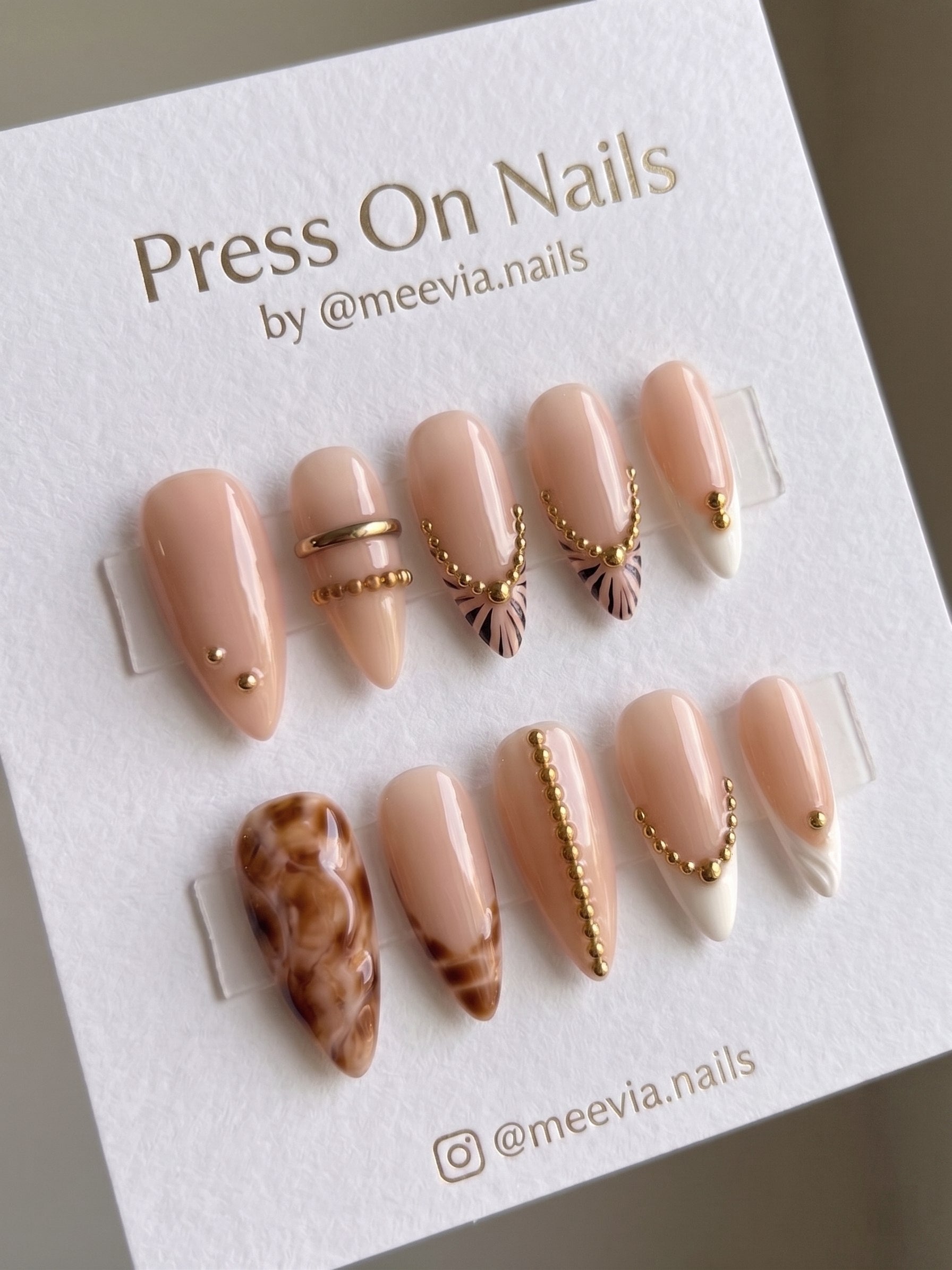 Almond-shaped nude press-on nails with gold bead chains and marble accents.