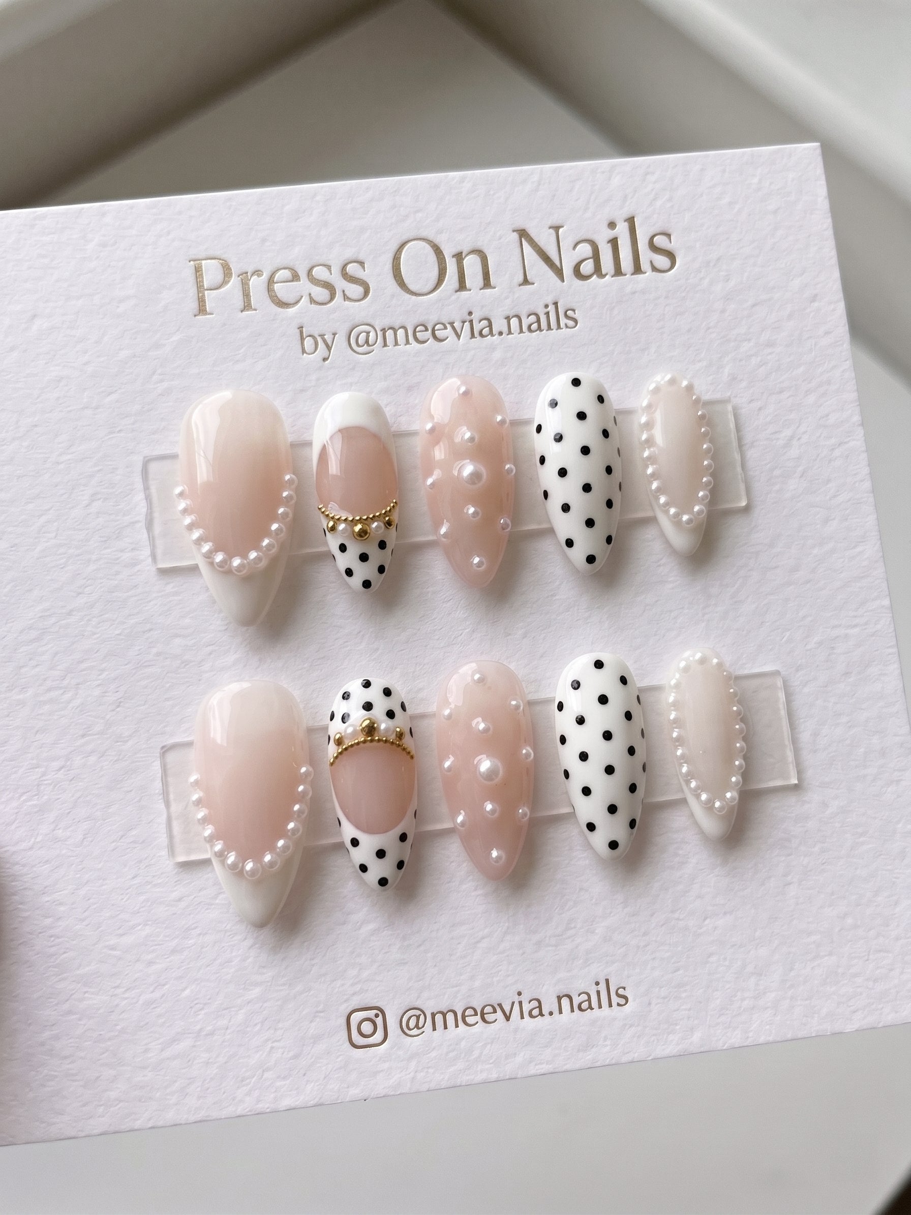 Almond-shaped press-on nails featuring white and nude colors with black polka dots, pearl borders, and gold bead accents as a complete nail set.