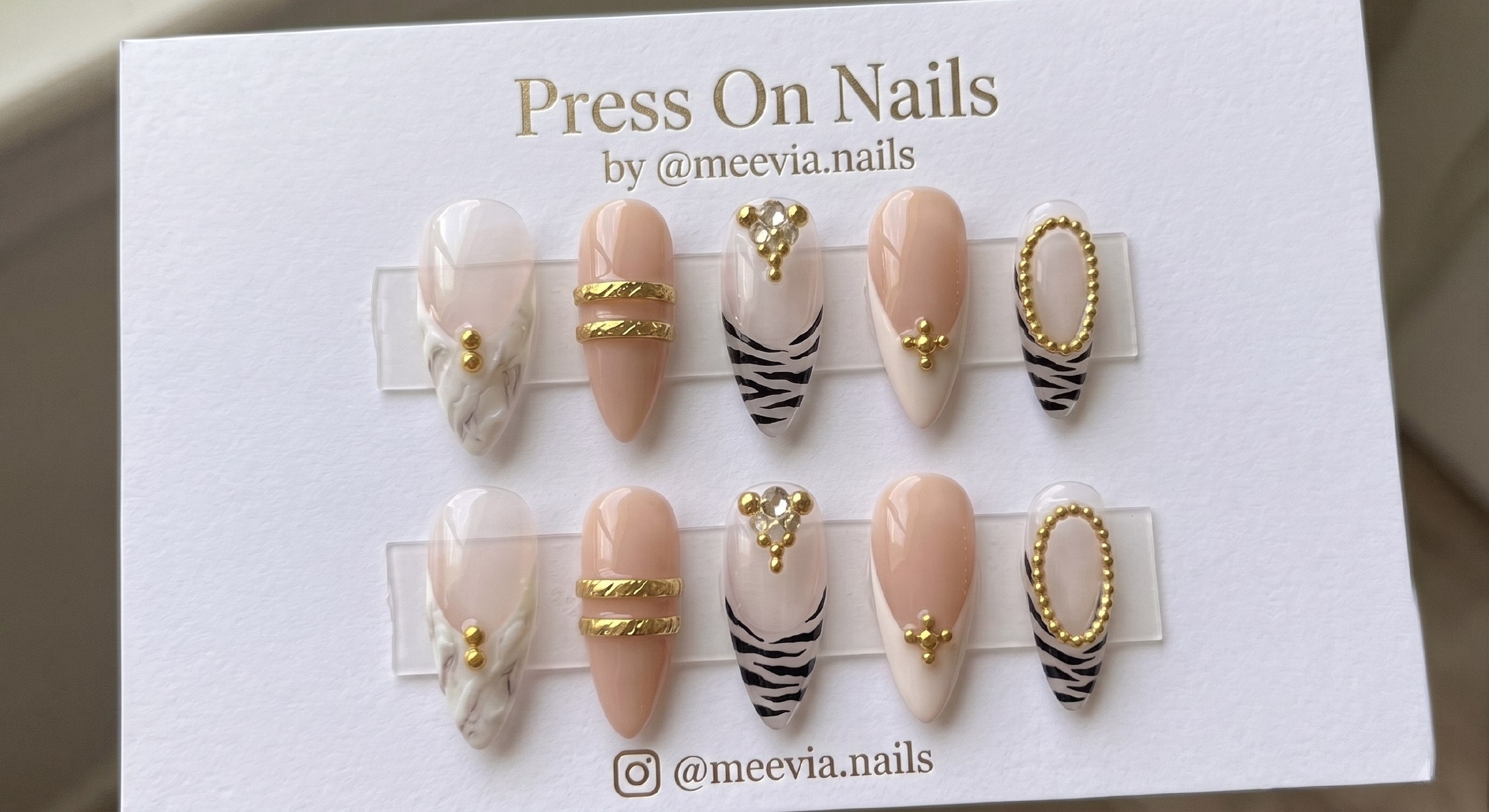 Almond-shaped nude and milky white press-on nails with black zebra stripes and 3D gold accents