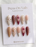 Stiletto-shaped press-on nails featuring burgundy croc print, 3D white floral designs, and gold bead embellishments.