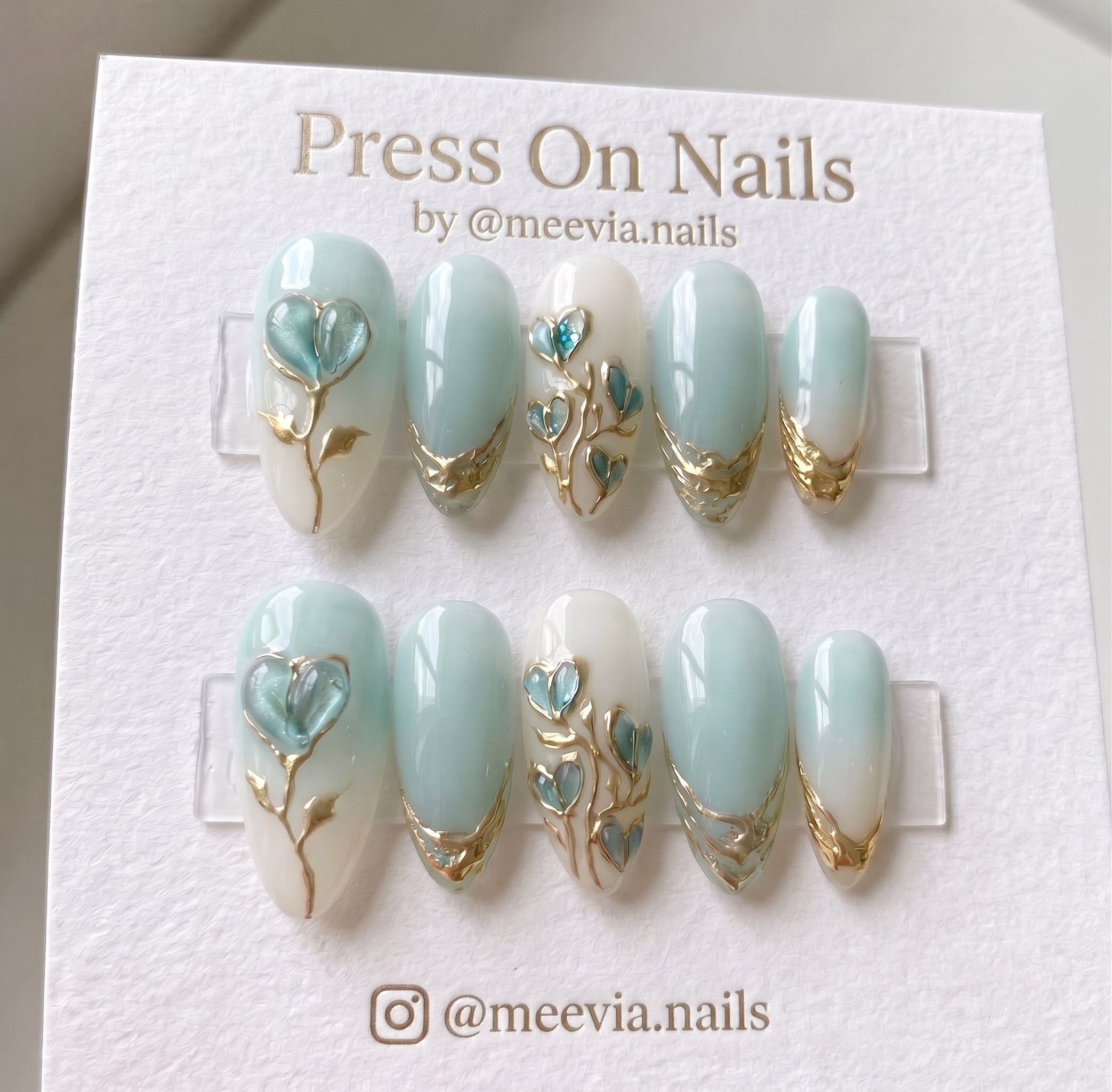 Almond-shaped mint blue and white press-on nails with 3D heart petals and gold metallic vine designs.