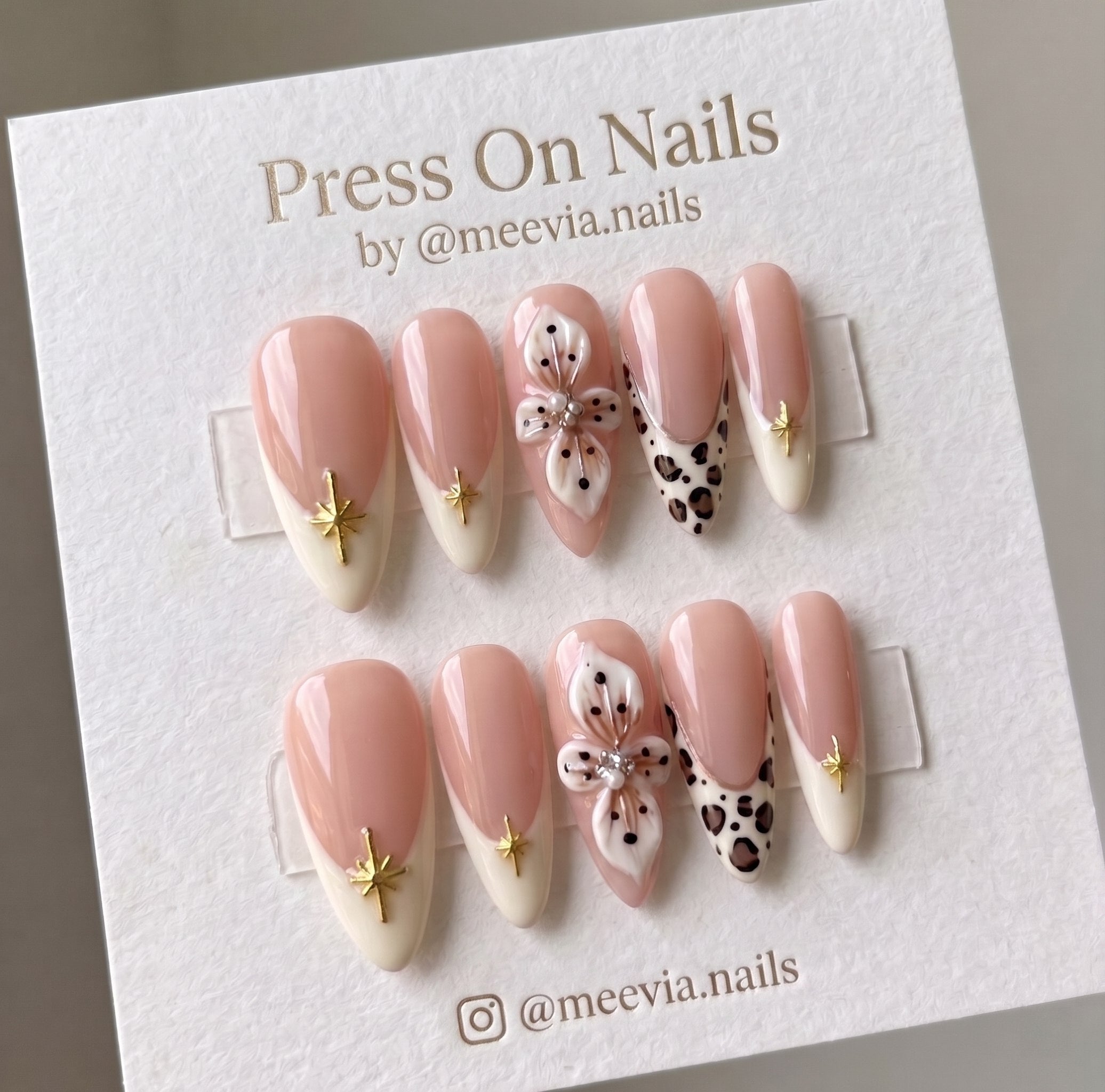 Almond-shaped nude press on nails with 3D white flowers, leopard print tips, and gold star charms.