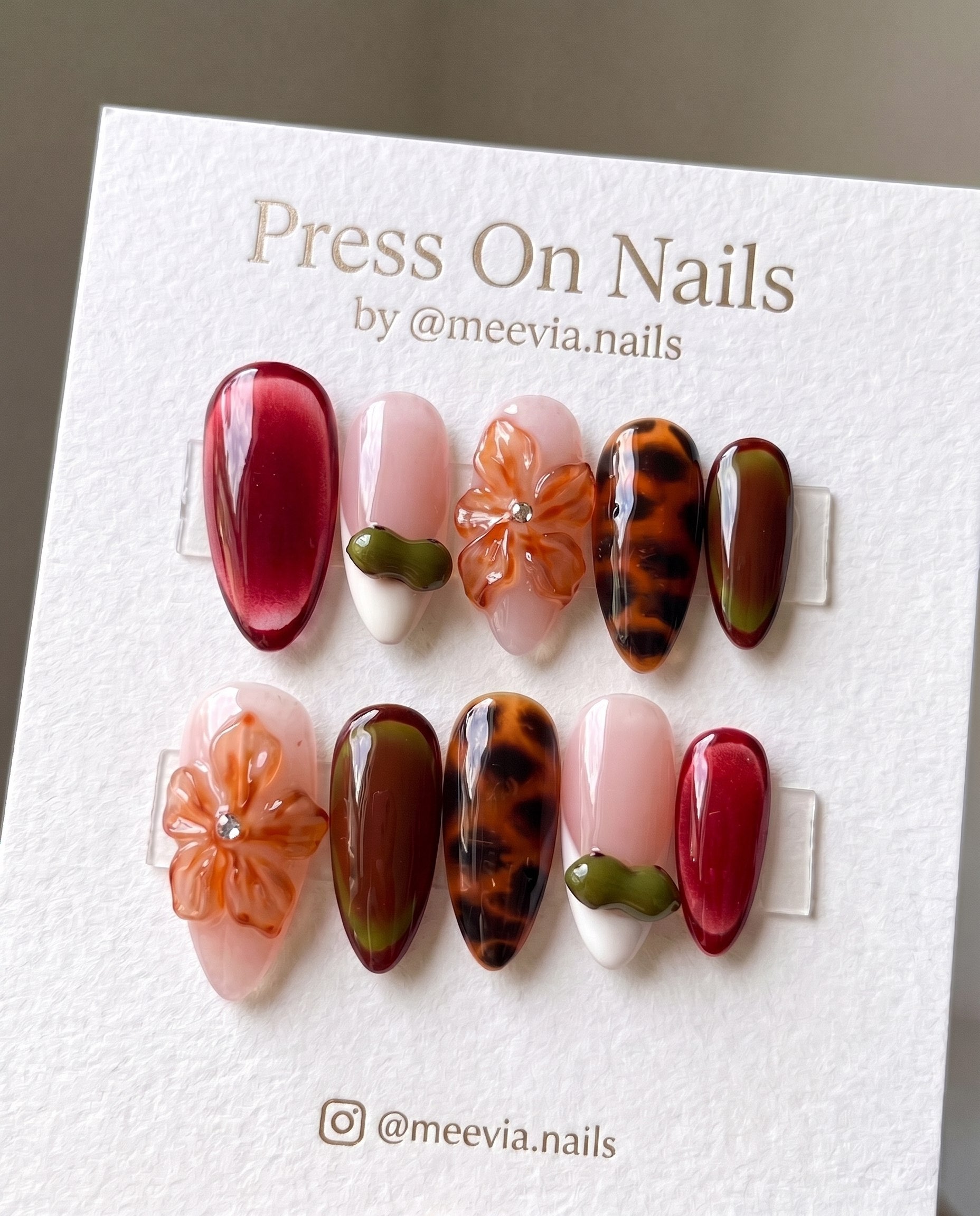 Almond-shaped press-on nails featuring 3D floral art, tortoiseshell patterns, and burgundy cat eye finish.