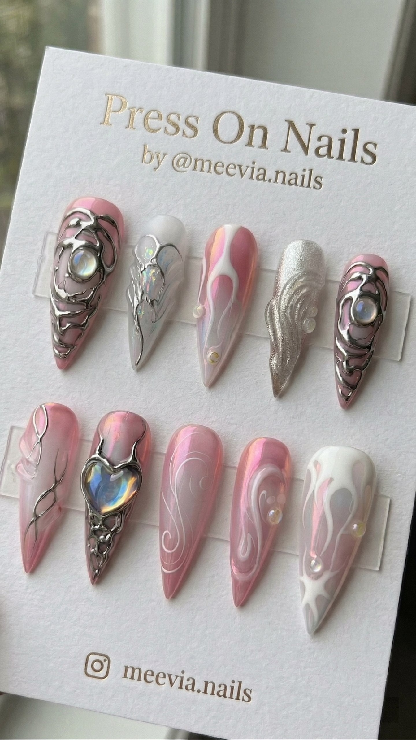 Long stiletto-shaped pink press-on nails with silver 3D chrome accents and iridescent heart gems.