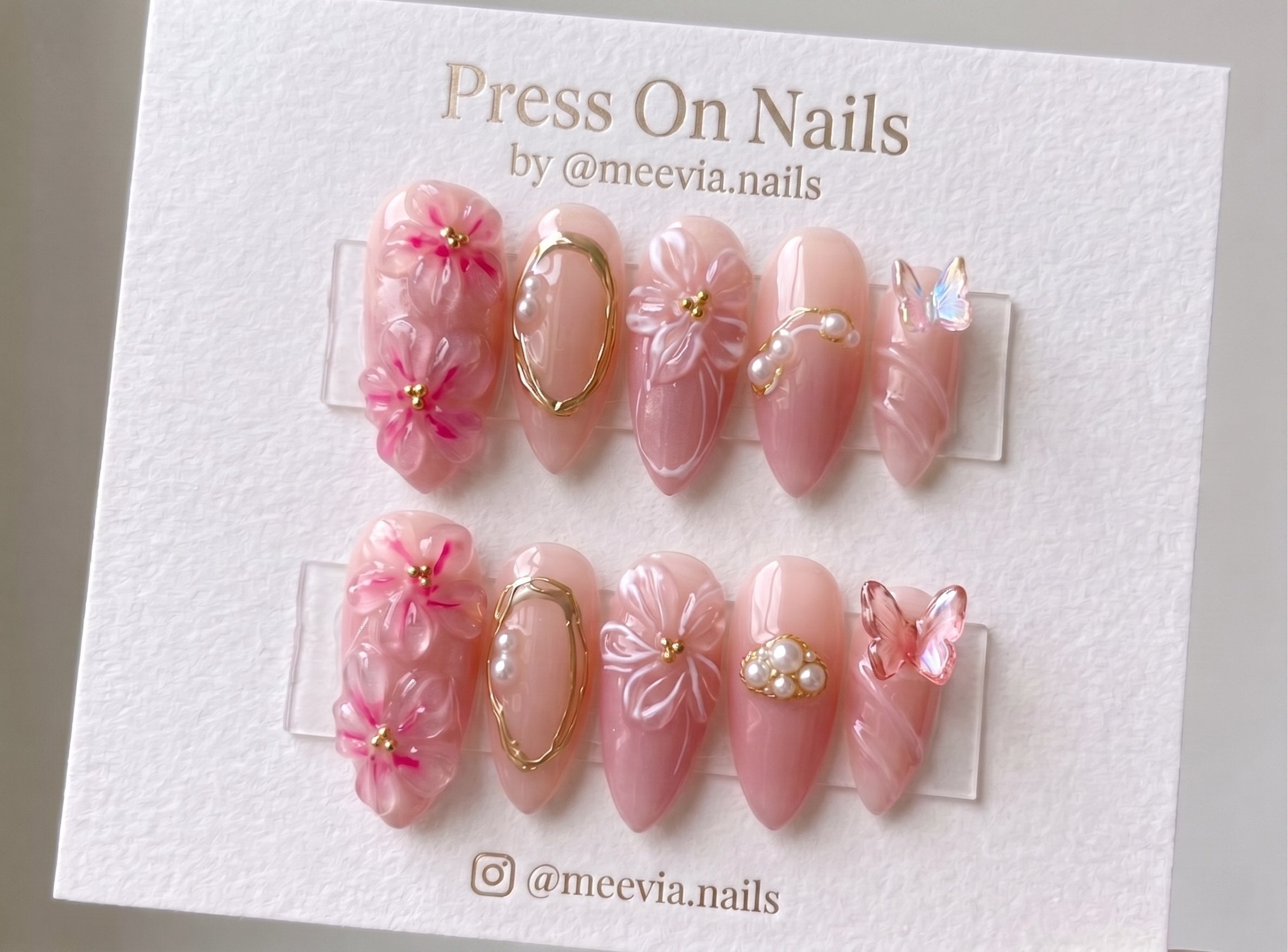 Almond-shaped pink press-on nails with 3D floral art, pearls, and butterfly accents