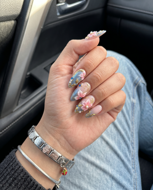Pink and grey gradient press-on nails featuring 3D floral art, gold metallic swirls, pearls, and blue rhinestones.