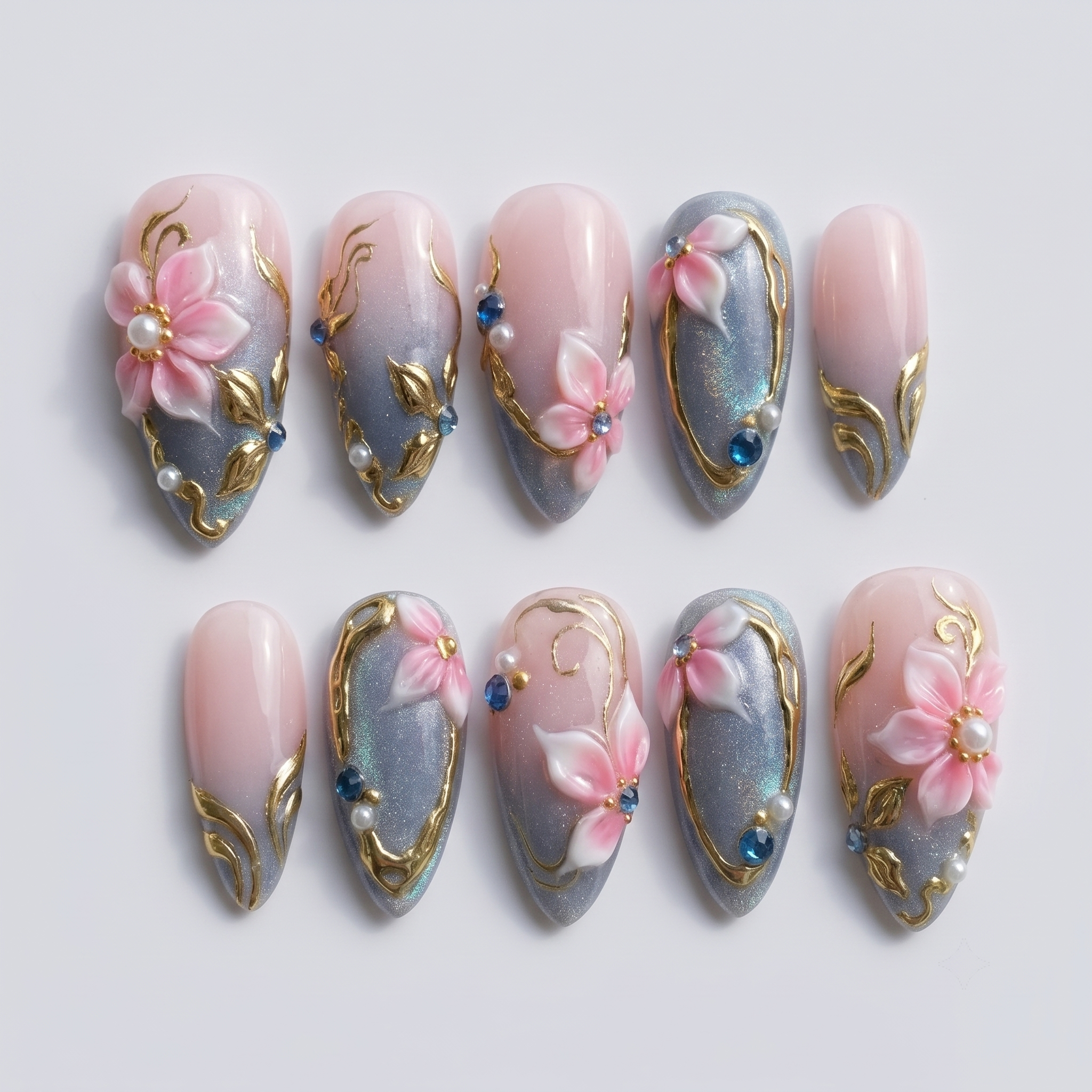 Pink and grey gradient press-on nails featuring 3D floral art, gold metallic swirls, pearls, and blue rhinestones.